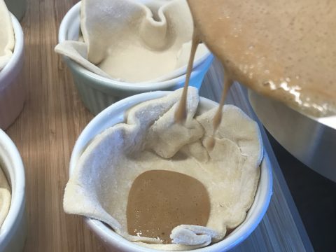 Eine cremige, braune Füllung wird in eine mit Tortenboden ausgelegte Auflaufform gegossen. Auf einer Holzoberfläche sind mehrere andere Auflaufformen mit demselben Boden zu sehen.