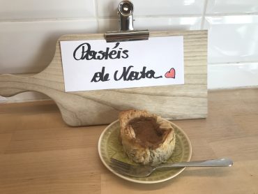 Ein portugiesisches Pastel de Nata liegt mit einer Gabel auf einem grünen Teller auf einem Holztisch. Dahinter liegt auf einem hölzernen Schneidebrett eine Notiz mit der Aufschrift „Pastéis de Nata“ und einem roten Herz. Im Hintergrund eine weiß geflieste Wand.