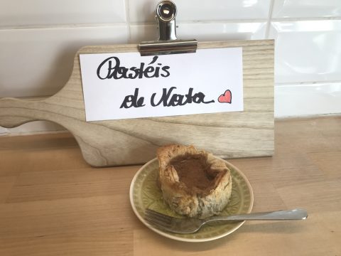 Ein portugiesisches Pastel de Nata liegt mit einer Gabel auf einem grünen Teller auf einem Holztisch. Dahinter liegt auf einem hölzernen Schneidebrett eine Notiz mit der Aufschrift „Pastéis de Nata“ und einem roten Herz. Im Hintergrund eine weiß geflieste Wand.