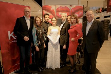 Eine Gruppe von sieben Personen, darunter eine Braut in einem weißen Kleid, steht zusammen in einem Einkaufszentrum. Sie posieren vor einem Hintergrund mit roten Lippenpaaren. Zwei Personen sind rot gekleidet, während die anderen dunkle Anzüge und Hemden tragen.