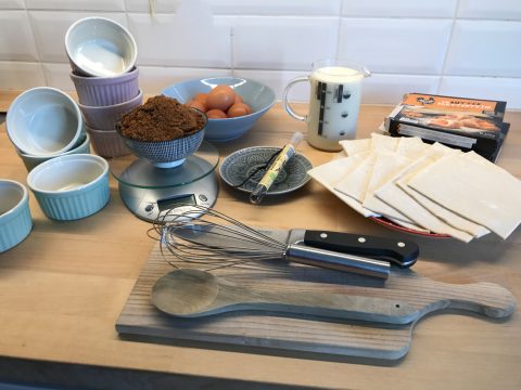 Eine Küchentheke mit Backzutaten und -utensilien, darunter brauner Zucker, Eier, Milch, Butter, Teigblätter, Auflaufförmchen, ein Schneebesen, ein Holzlöffel und ein Spatel. Alle Gegenstände sind ordentlich vor einer weiß gefliesten Wand angeordnet.