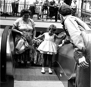 Ein junges Mädchen im Kleid betritt mit Hilfe eine Rolltreppe, während eine Frau mit Taschen in der Hand daneben steht. Im Hintergrund schauen andere Leute zu. Schwarzweißbild.