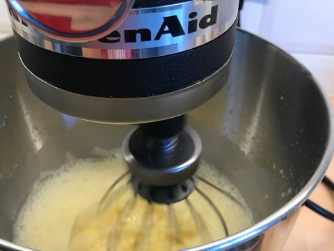 Eine Nahaufnahme einer Küchenmaschine, die Zutaten in einer Edelstahlschüssel vermengt. Die Mischung darin sieht leicht und schaumig aus, was darauf hindeutet, dass sie gerade dabei ist, einen Teig zu schlagen oder zu mischen. Die Maschine trägt die Marke „KitchenAid“.