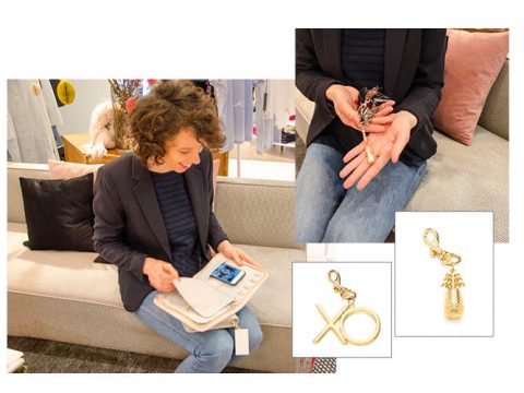Eine Person mit lockigem Haar sitzt auf einem Sofa und zeichnet in einem Notizbuch. In der Nähe sind Nahaufnahmen ihrer Hände zu sehen, die Schmuck und zwei Anhänger halten, einer in Form von „XO“ und der andere in Form einer Ananas, beide mit Verschluss.