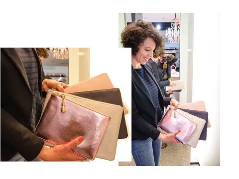 Eine Person hält in einem Geschäft mehrere farbige Clutches, darunter Metallic-Pink, Beige und Schwarz. Die Person lächelt und trägt einen schwarzen Blazer und ein gestreiftes Hemd. Im Hintergrund ist ein Kronleuchter zu sehen.