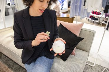Eine Person sitzt in einem Geschäft auf einem Sofa. Sie trägt einen schwarzen Blazer und Jeans und hält einen kleinen verpackten Artikel in der Hand. Im Hintergrund sind Kleiderständer und ein Bildschirm zu sehen.