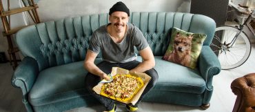 Eine Person mit Beanie und legerer Kleidung sitzt auf einem blaugrünen Sofa und hält eine frisch gebackene Pizza auf Backpapier in den Händen. Daneben liegt ein dekoratives Kissen mit einem Fuchsmotiv und im Hintergrund ist teilweise ein Fahrrad zu sehen.