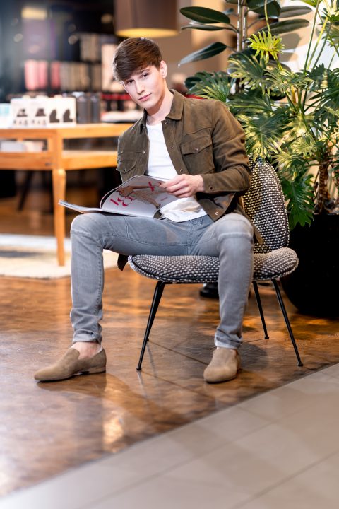 Eine Person sitzt in einem modernen Innenraum auf einem Stuhl und liest eine Zeitschrift. Sie trägt eine braune Jacke, ein weißes Hemd, graue Jeans und beige Schuhe. Im Hintergrund sind große grüne Pflanzen und Regale zu sehen.