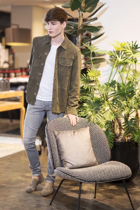 Eine Person mit kurzen braunen Haaren, die eine grüne Jacke, ein weißes Hemd und graue Jeans trägt, steht neben einem gepolsterten Stuhl. Im Hintergrund sind eine Pflanze und eine Arbeitsplatte zu sehen.