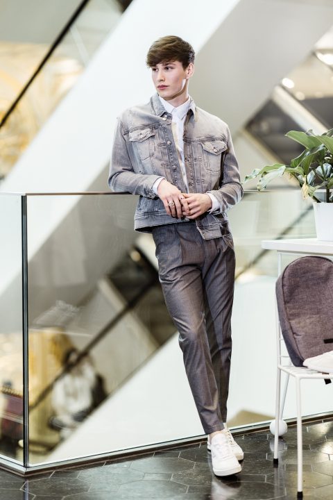 Ein junger Mann lehnt lässig an einem Glasgeländer in einem modernen Gebäude. Er trägt eine Jeansjacke, ein weißes Hemd und graue Hosen sowie weiße Turnschuhe. Auf einem Tisch in der Nähe ist eine kleine Pflanze zu sehen. Geometrische Hintergründe verleihen dem Ganzen eine stilvolle Note.