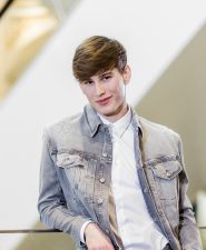Eine Person, die eine helle Jeansjacke über einem weißen Hemd trägt, steht im Innenbereich, leicht nach vorne geneigt und hat die Hände in den Taschen. Der Hintergrund ist leicht verschwommen und weist diagonale Linien und einen warmen Lichtschein auf.
