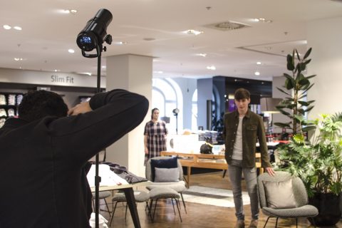 Ein Fotograf macht ein Foto von einem Mann, der lässig in einem modernen Bekleidungsgeschäft posiert. Im Hintergrund steht eine Frau. Das Geschäft ist gut beleuchtet, die Kleidung ist ausgestellt und es gibt eine Vielzahl von Möbeln und Pflanzen.