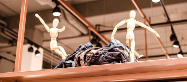 Zwei hölzerne Schaufensterpuppen stehen verspielt auf einem Ladenregal mit einem Stapel gefalteter Jeans und Gürtel. Die Kulisse scheint ein Bekleidungsgeschäft zu sein, im Hintergrund sind Lichter und Deckenleuchten zu sehen.