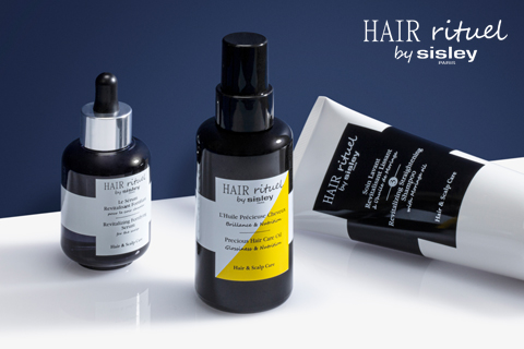 Eine Kollektion von Haarpflegeprodukten der Marke „HAIR rituel by sisley“ auf einer Oberfläche. Das Set enthält eine Tropfflasche, eine Sprühflasche und eine Tube, alle mit der Aufschrift „Haar- und Kopfhautpflege“ und einem dunklen Hintergrund.