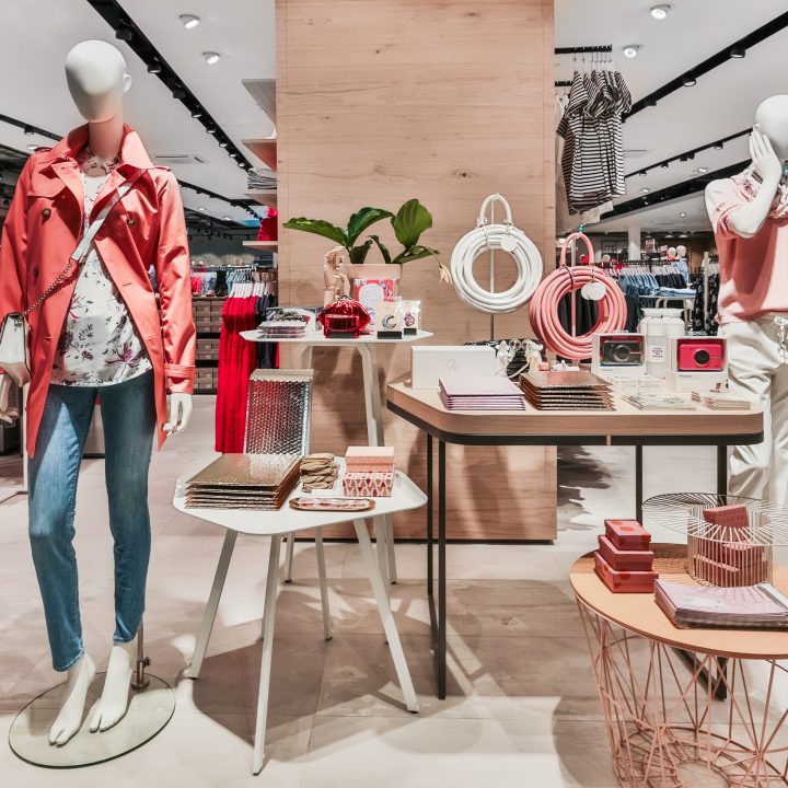 Ein stilvolles Schaufenster in einem Bekleidungsgeschäft mit Schaufensterpuppen in legerer Kleidung. Die linke Schaufensterpuppe trägt eine rote Jacke, eine bedruckte Bluse und Jeans, während die rechte Schaufensterpuppe ein rosa Hemd und weiße Hosen trägt. Accessoires sind auf den Tischen daneben angeordnet.