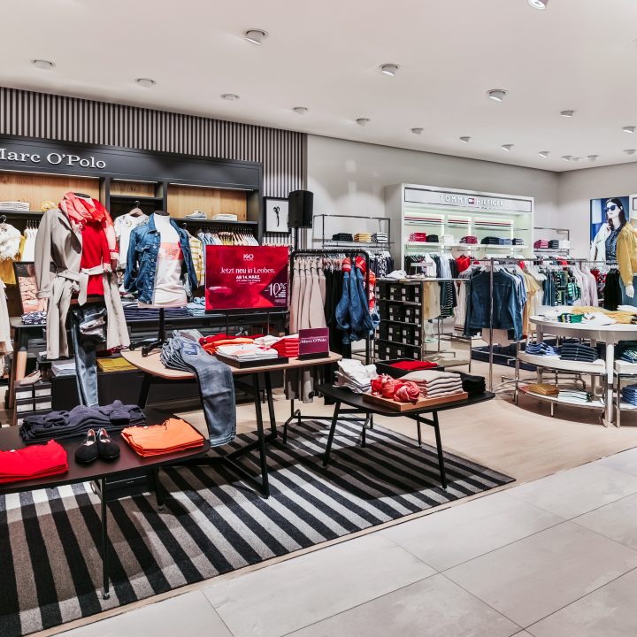 Ein modernes Interieur eines Bekleidungsgeschäfts mit ordentlich angeordneten Kleiderreihen auf Ständern und Tischen. Helles Licht hebt verschiedene Kleidungsstücke hervor, darunter Hemden, Jacken und Jeans. Markenschilder für Mare O’Polo und Tommy Hilfiger sind sichtbar.