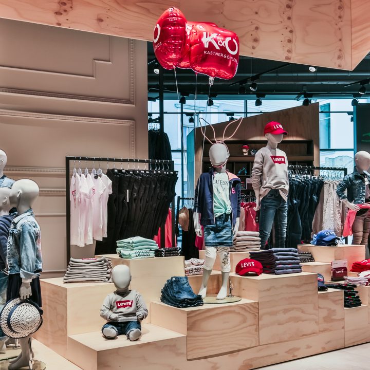 Schaufenster eines Kinderbekleidungsgeschäfts mit Schaufensterpuppen in legerer Kleidung, darunter Jeansjacken und Levi's-Hemden. Darüber schweben rote und weiße Luftballons. Die Regale sind voll mit gefalteter Kleidung und das Geschäft ist hell erleuchtet.