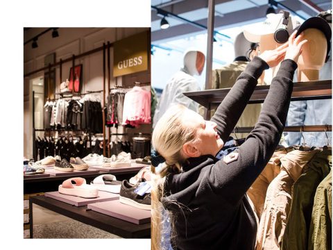 Eine Frau ordnet Hüte auf einem Display in einem Bekleidungsgeschäft an. Im Hintergrund sind auf Regalen Turnschuhe und Kleiderständer mit Kleidung, darunter Jacken und Kapuzenpullover, ausgestellt. Im Laden sind verschiedene Marken zu sehen.