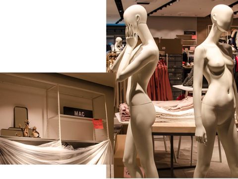 Zwei Schaufensterpuppen stehen neben einem Tisch in einem Bekleidungsgeschäft; eine posiert nachdenklich. In der Nähe sind Regale mit Artikeln und Stoffen zu sehen, die darüber drapiert sind. Die Szene scheint Teil einer Verkaufsausstellung zu sein.