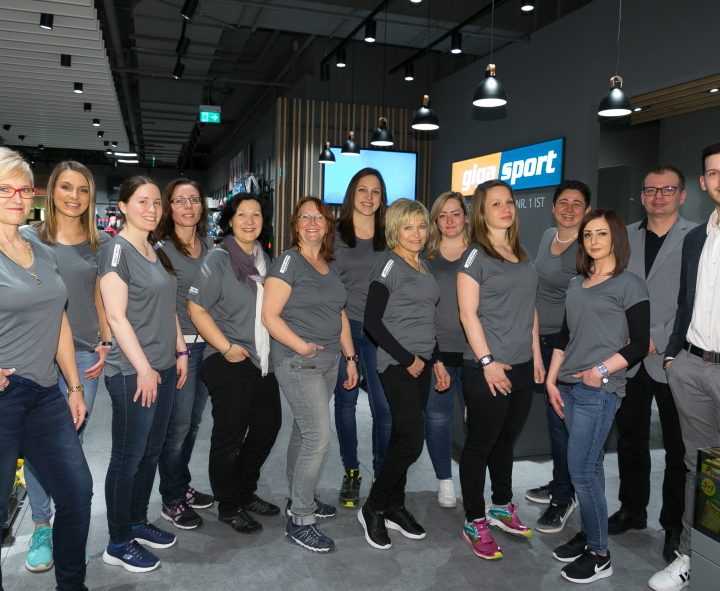 Eine Gruppe von dreizehn Personen, hauptsächlich Frauen, steht in einem Sportartikelgeschäft zusammen und trägt passende graue Hemden. Im Hintergrund sind das Innere des Ladens und verschiedene Sportartikel zu sehen.
