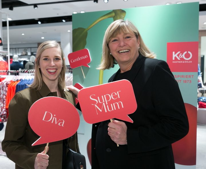 Zwei Frauen lächeln und halten rote Sprechblasenschilder. Auf einem Schild steht „Diva“, auf dem anderen „Supermama“. Sie stehen in einem Geschäft, im Hintergrund sind Kleidung und Marken zu sehen.