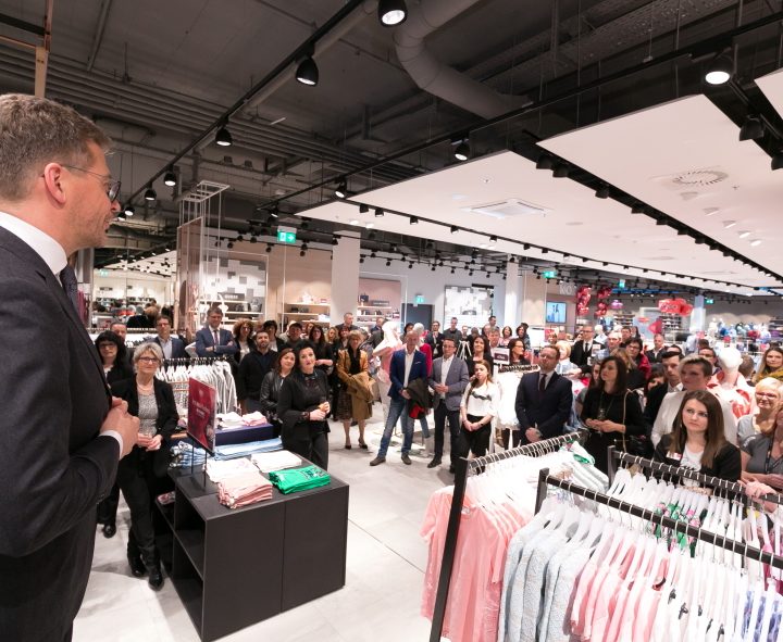 Ein Mann im Anzug spricht zu einer großen Gruppe von Menschen, die sich in einem Bekleidungsgeschäft versammelt haben. Man sieht Kleiderständer mit gefalteter und hängender Kleidung. Die Anwesenden hören aufmerksam zu, trotz der hellen Beleuchtung und der modernen Einrichtung des Ladens.