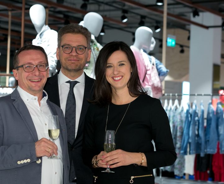 Drei Personen stehen lächelnd in einem Bekleidungsgeschäft und halten Champagnergläser in den Händen. Im Hintergrund sind Schaufensterpuppen in Freizeitkleidung und Kleiderständer mit Kleidung zu sehen. Die Atmosphäre ist festlich und fröhlich.