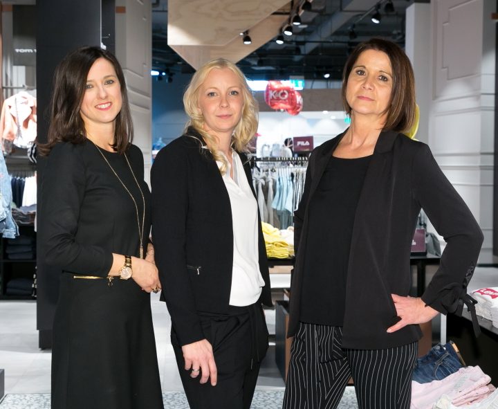 Drei Frauen posieren selbstbewusst in einem schicken Bekleidungsgeschäft. Sie stehen zwischen Kleiderständern mit ordentlich präsentierter Kleidung, im Hintergrund sind legere und formelle Kleidung zu sehen. Das Ambiente ist modern und gut beleuchtet.