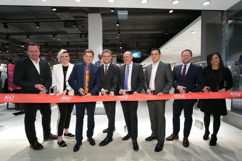 Eine Gruppe von acht Personen steht in einem Geschäft und schneidet mit einer Schere ein rotes Band mit der Aufschrift „KO“ durch. Damit wird die Eröffnung eines neuen Ladenlokals markiert. Sie tragen Business-Kleidung und das Ambiente ist hell und modern eingerichtet.