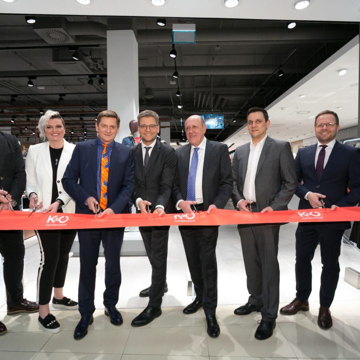 Eine Gruppe von acht Personen steht in einem Geschäft und schneidet mit einer Schere ein rotes Band mit der Aufschrift „KO“ durch. Damit wird die Eröffnung eines neuen Ladenlokals markiert. Sie tragen Business-Kleidung und das Ambiente ist hell und modern eingerichtet.