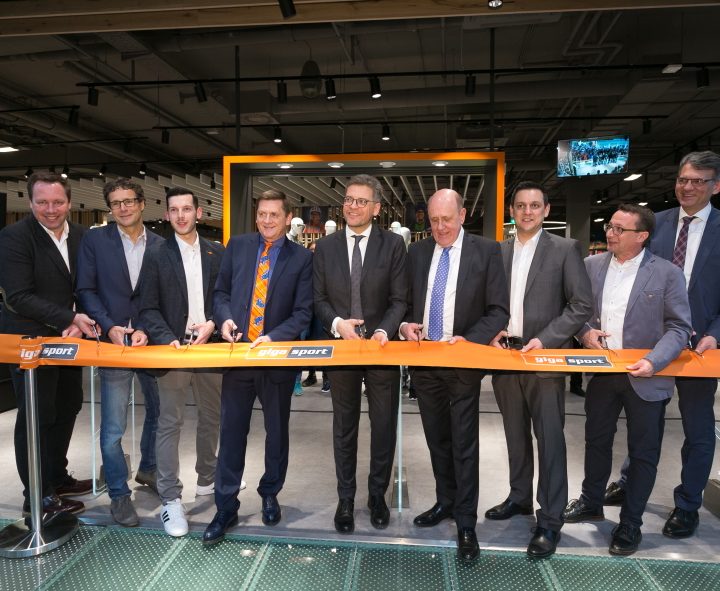 Neun Männer in Businesskleidung schneiden bei einer Indoor-Veranstaltung, möglicherweise einer Geschäftseröffnung, ein orangefarbenes Band durch. Sie stehen lächelnd in einer Reihe, jeder hält eine Schere in der Hand, im Hintergrund eine moderne Innenkulisse.