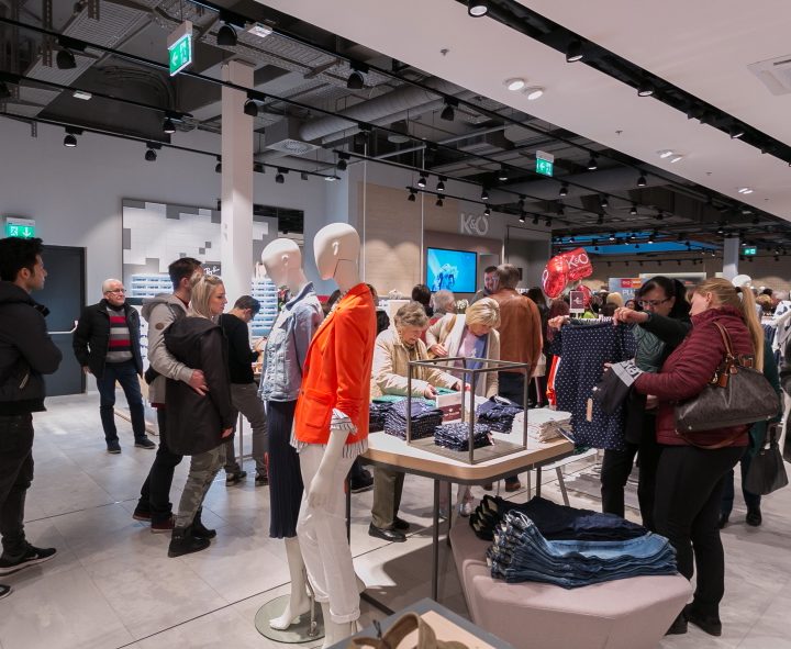 Kunden stöbern in einem geschäftigen Einzelhandelsgeschäft durch Kleiderauslagen. Schaufensterpuppen präsentieren die Kleidungsstücke neben einem Tisch mit gefalteter Kleidung. Käufer, darunter einige, die die Artikel genau unter die Lupe nehmen, bewegen sich in dem gut beleuchteten Raum mit modernem Dekor umher.
