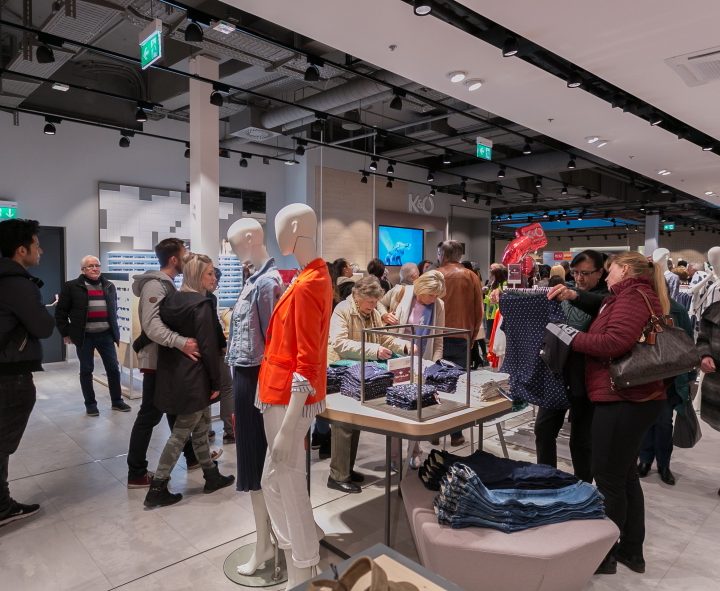 Menschen kaufen in einem geschäftigen Bekleidungsgeschäft ein. Schaufensterpuppen präsentieren verschiedene Outfits, darunter eine leuchtend orangefarbene Jacke. Käufer stöbern in Regalen und auf Tischen voller Kleidung. Das Geschäft hat ein modernes Innendesign mit Deckenbeleuchtung.
