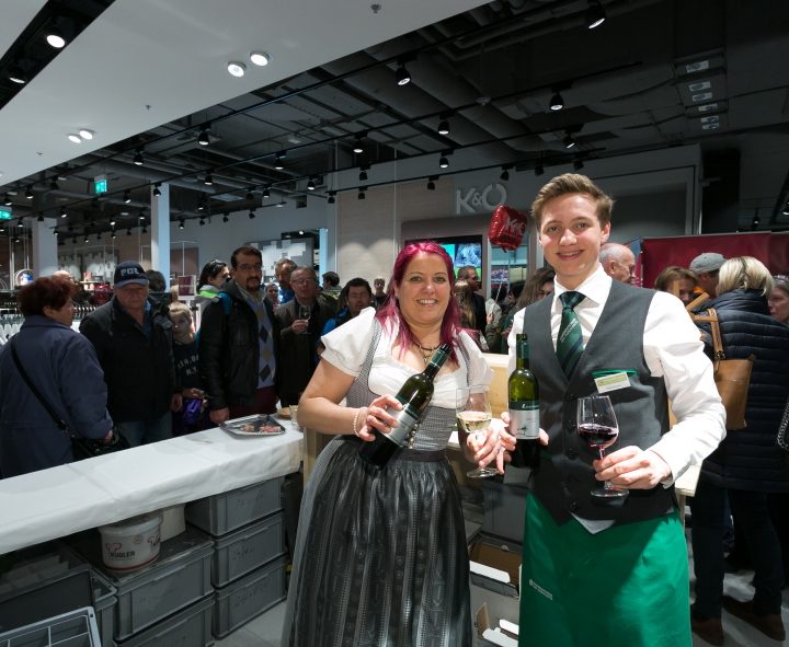 Eine Frau und ein Mann servieren Wein in einem geschäftigen Geschäft. Die Frau trägt ein Dirndl und hält zwei Weinflaschen; der Mann ist weiß gekleidet, trägt eine grüne Schürze und hält eine Weinflasche und ein Glas. Im Hintergrund hat sich eine Menschenmenge versammelt.