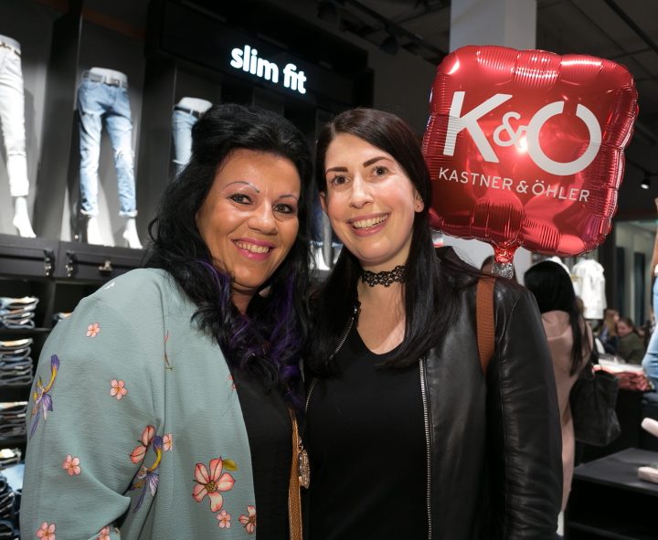Zwei Frauen lächeln in einem Bekleidungsgeschäft. Eine trägt einen geblümten Schal, die andere eine schwarze Jacke. Sie halten einen roten Luftballon mit der Aufschrift „K&Ö Kastner & Öhler“. Im Hintergrund sind Jeans ausgestellt.