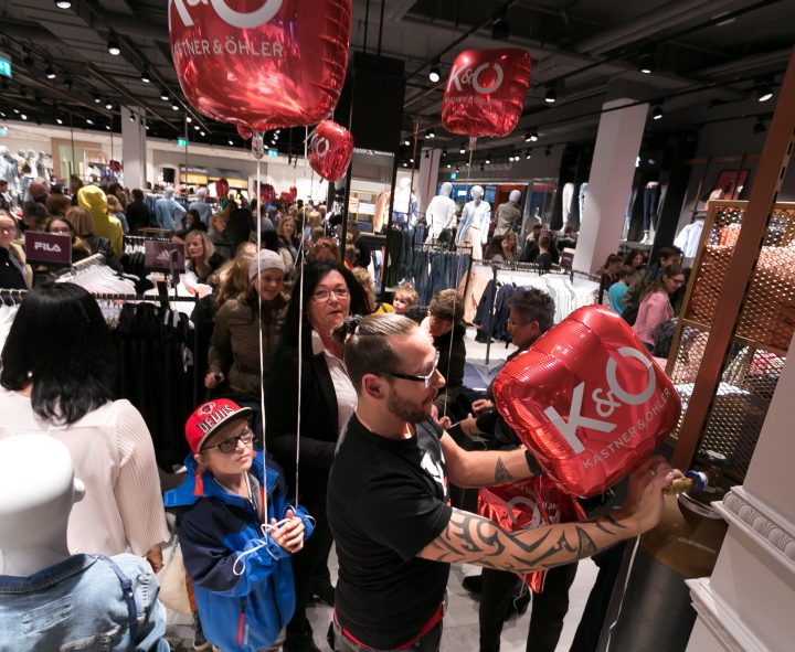 Ein überfüllter Laden mit Menschen, die einkaufen, darunter ein Mann, der rote Luftballons mit dem Aufdruck „K&O“ verteilt. Ein Kind in einer blauen Jacke und Mütze steht daneben und schaut sich die Luftballons an. Im Hintergrund sind Schaufensterpuppen zu sehen, die Kleidung präsentieren.