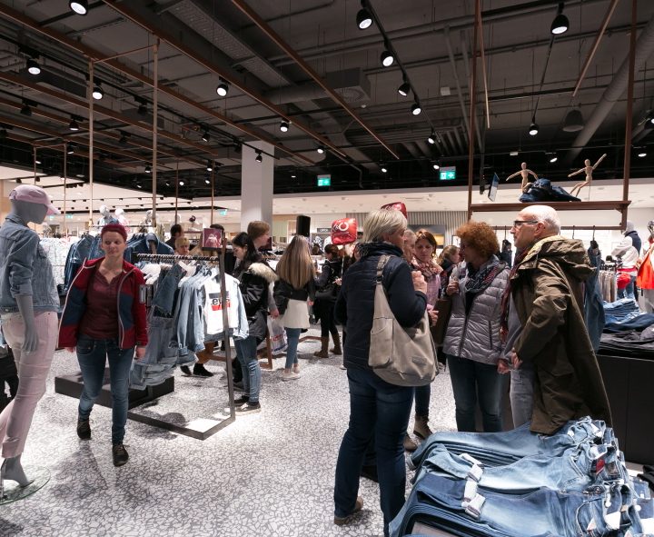 Menschen gehen in einem Bekleidungsgeschäft einkaufen. Schaufensterpuppen und Kleiderständer mit verschiedenen Kleidungsstücken sind zu sehen. Das Geschäft ist hell beleuchtet und modern gestaltet. Mehrere Kunden stöbern und unterhalten sich in den Gängen.