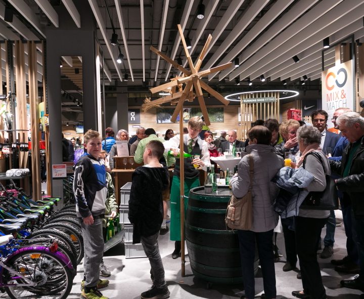 Eine gut besuchte Veranstaltung im Innenbereich, bei der sich die Leute um einen Holztisch herum tummeln. Auf der linken Seite stehen Fahrräder, und an den Wänden hängen bunte Waren. Die Decke ist modern beleuchtet. Die Leute tragen legere und halbformelle Kleidung.