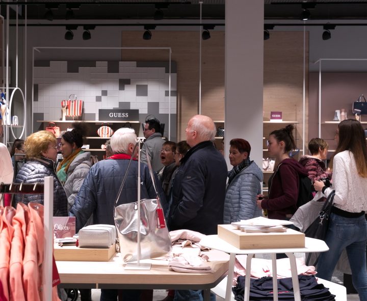 Eine Gruppe von Menschen geht in einem Bekleidungsgeschäft einkaufen. Sie stöbern in Kleiderständern mit Kleidung und Accessoires. Das Geschäft ist hell beleuchtet und modern ausgestattet. Mehrere Käufer tragen Jacken und konzentrieren sich auf die Artikel vor ihnen.