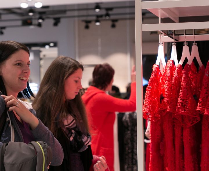 Zwei Frauen kaufen in einem Bekleidungsgeschäft ein. Sie stöbern neben einem Ständer mit roten Spitzenkleidern. Eine Frau hält eine Jacke und eine Tasche, während die andere sich die Kleider ansieht. Das Geschäft ist hell beleuchtet und modern gestaltet.