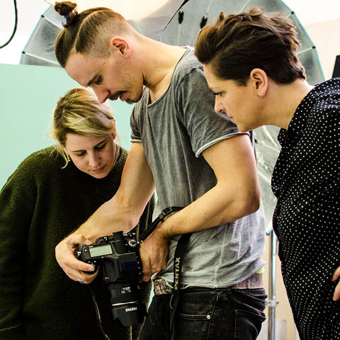 Drei Personen prüfen während eines Fotoshootings Bilder auf einer Digitalkamera. Die Person in der Mitte hält die Kamera, während die anderen beiden zuschauen. Im Hintergrund ist eine Studiobeleuchtungsausrüstung zu sehen.