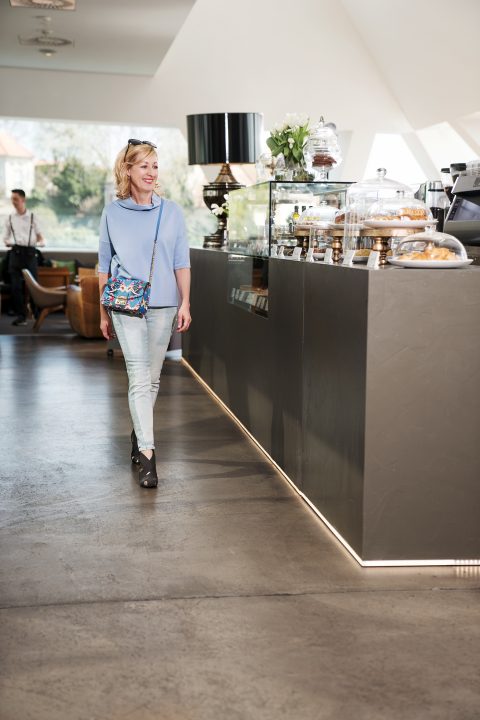 Eine Person in hellblauem Pullover und Jeans betritt ein modernes Café. Sie lächelt und trägt eine bunte Umhängetasche. Auf der Theke rechts von ihr sind unter Glaskuppeln verschiedene Backwaren und Kuchen ausgestellt.