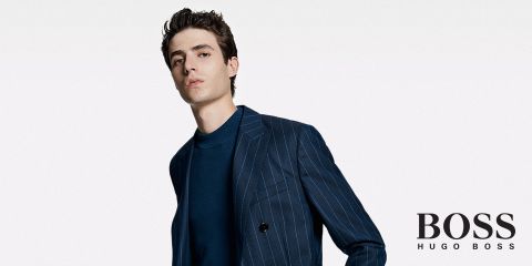 Eine Person, die einen dunklen Nadelstreifenanzug über einem blauen Pullover trägt, steht vor einem schlichten weißen Hintergrund. In der unteren rechten Ecke ist das Logo „BOSS Hugo Boss“ zu sehen.