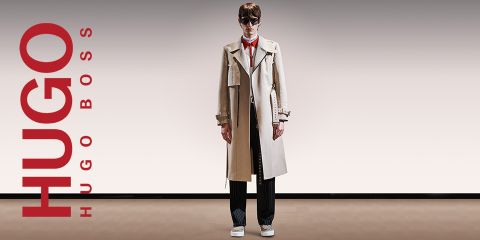 Eine Person in einem beigen Trenchcoat, einer schwarzen Hose und weißen Schuhen steht vor einem Hintergrund mit Farbverlauf. Links ist vertikal in Rot der Schriftzug „HUGO BOSS“ zu lesen.