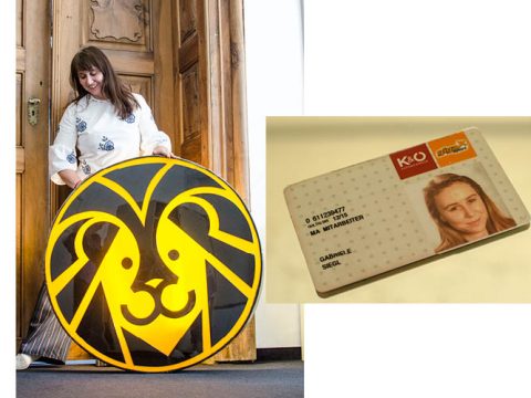 Eine Frau lächelt und hält ein großes, rundes Schild mit einem stilisierten gelb-schwarzen Löwengesicht. Neben ihr liegt ein Ausweis, auf dem ein Teilprofilbild und Identifikationsdaten einer Frau zu sehen sind.