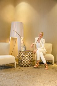 Eine Person im weißen Anzug sitzt auf einem stylischen Sessel in einem modernen Raum mit beigen Wänden. Daneben steht ein dekorativer Beistelltisch mit einer Vase und einer großen Stehlampe. Die Person lächelt und trägt braune Sandalen.