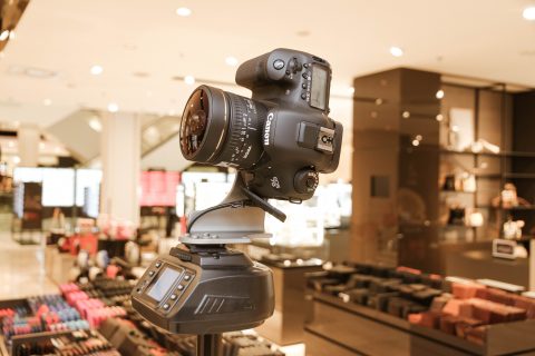 Eine professionelle DSLR-Kamera auf einem Stativ in einem hell erleuchteten Geschäft mit Regalen voller Waren, darunter Taschen und Accessoires, im Hintergrund.