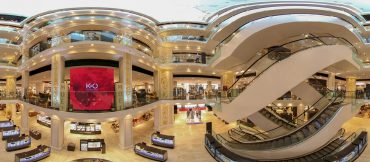 Ein Panoramablick auf das Innere eines luxuriösen mehrstöckigen Einkaufszentrums. Zu sehen sind mehrere Stockwerke mit Glasgeländern, eine Rolltreppe, ein großer Videobildschirm mit einem roten Bild und verschiedene Einzelhandelsauslagen im Erdgeschoss.