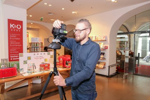 Ein Mann justiert in einem Geschäft eine auf einem Stativ montierte Kamera. Er trägt eine Brille und ein dunkles Langarmhemd. Hinter ihm sind Regale mit Küchenartikeln und ein Schild mit Produktinformationen. Das Geschäft ist modern gestaltet und hat gewölbte Glastüren.