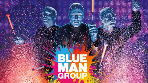 Drei Darsteller mit blauen Gesichtern und dunkler Kleidung halten bunte Trommelstöcke. Sie stehen vor einem lebhaften, mit Farbe bespritzten Hintergrund. Der Text lautet „Blue Man Group“ mit einem bunten Farbspritzer-Design darunter.